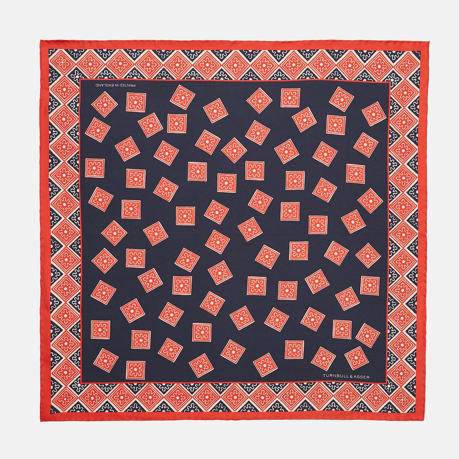 View 1 of product The Navy and Cherry Red Tiled Floral Silk Pocket Square by Turnbull & Asser features a blue background with red and white geometric floral patterns, a decorative border, and the brand's name in one corner.