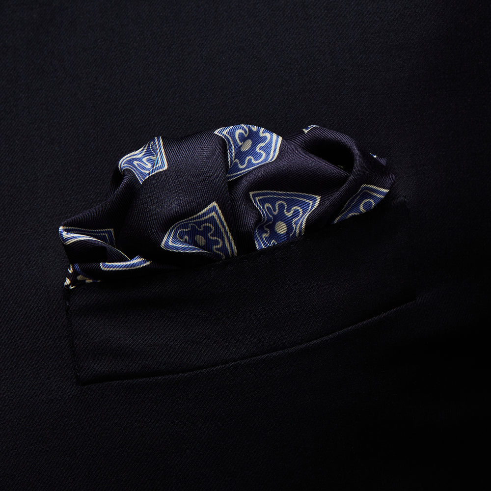 The Navy and Sky Blue Tiled Floral Silk Pocket Square by Turnbull & Asser features a geometric floral pattern in blue and white, a detailed border, and the brand's logo in the bottom right corner.