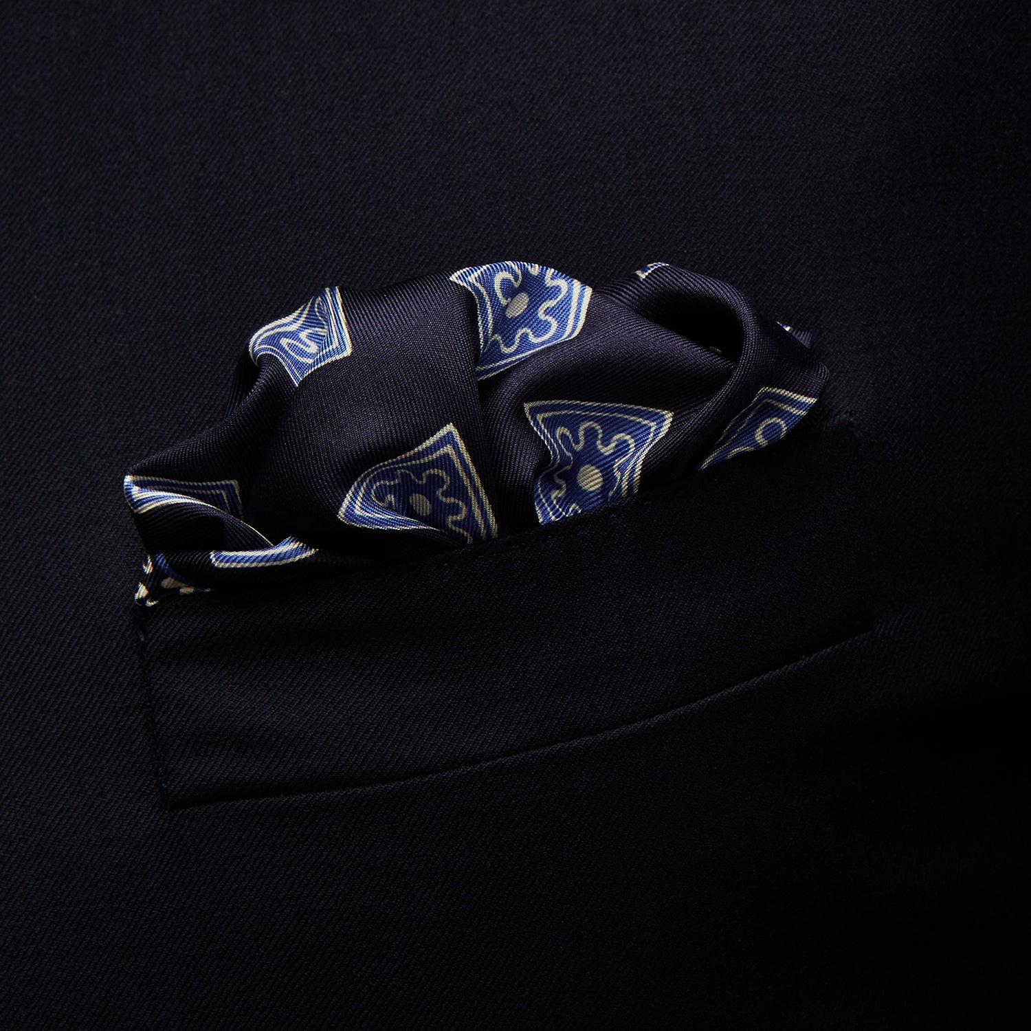 View 3 of product The Navy and Sky Blue Tiled Floral Silk Pocket Square by Turnbull & Asser features a geometric floral pattern in blue and white, a detailed border, and the brand's logo in the bottom right corner.