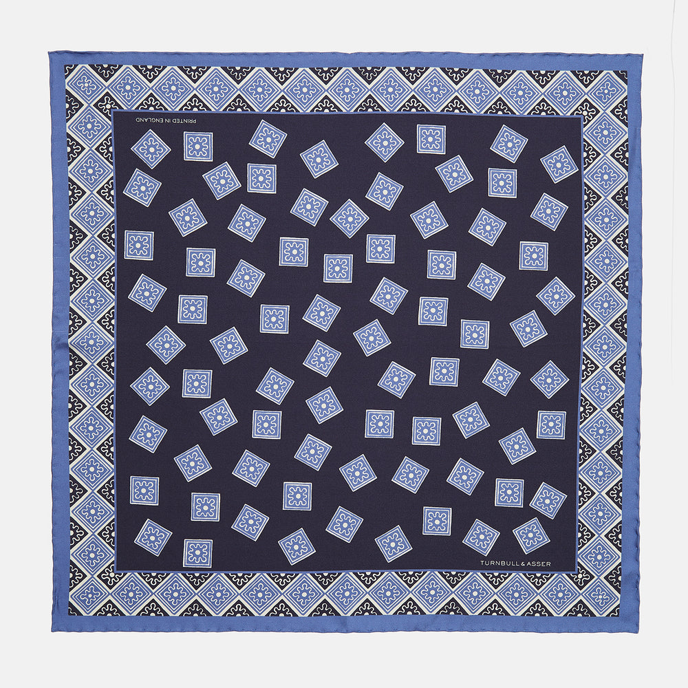 The Navy and Sky Blue Tiled Floral Silk Pocket Square by Turnbull & Asser features a geometric floral pattern in blue and white, a detailed border, and the brand's logo in the bottom right corner.