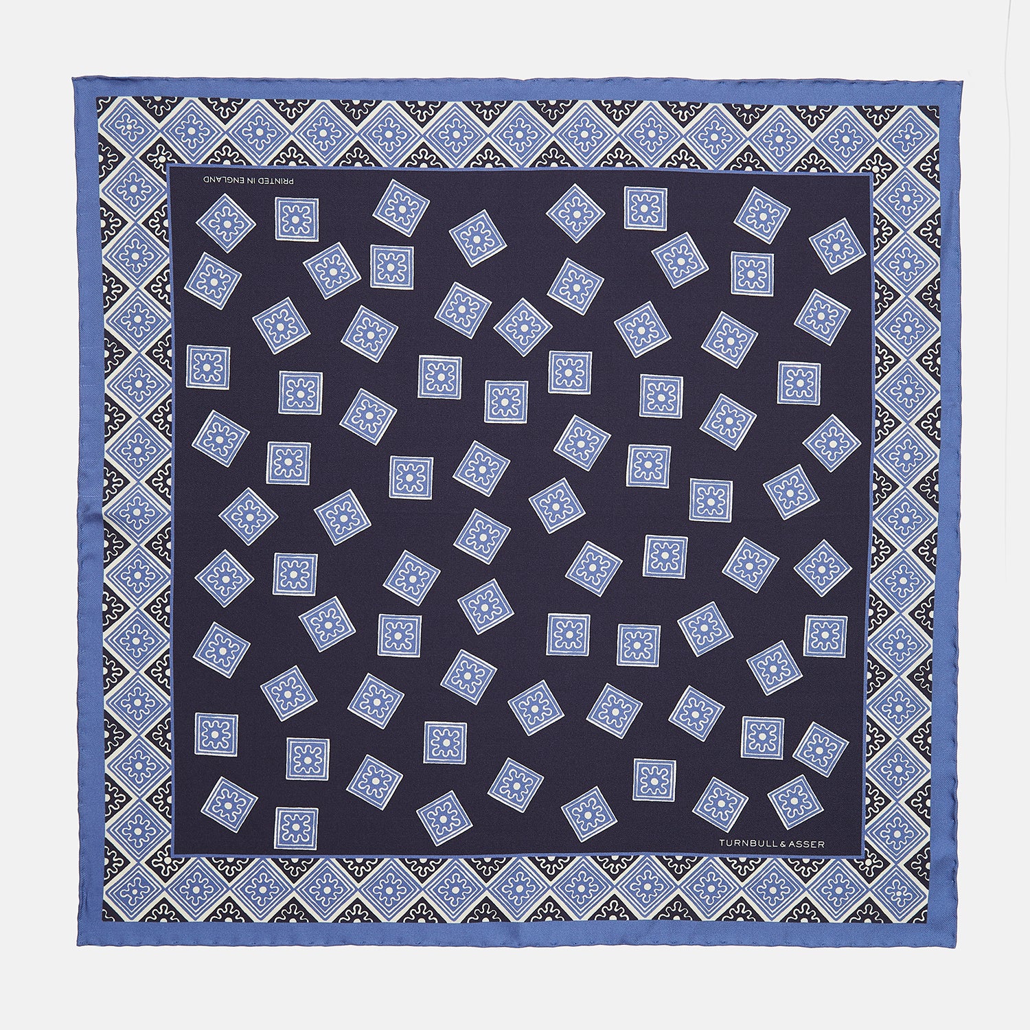 View 1 of product The Navy and Sky Blue Tiled Floral Silk Pocket Square by Turnbull & Asser features a geometric floral pattern in blue and white, a detailed border, and the brand's logo in the bottom right corner.