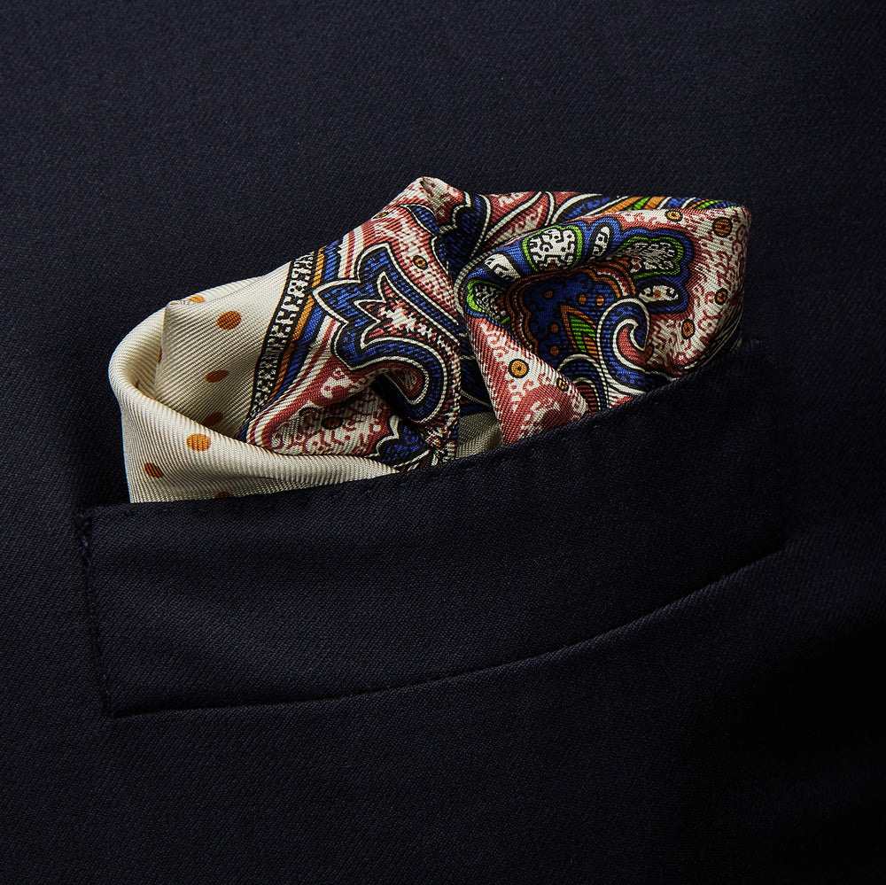 The Cream Paisley Polka Dot Silk Pocket Square by Turnbull & Asser features hand-rolled edges, a detailed blue, red, and yellow paisley design with central orange dots, and the brand's name printed in one corner.