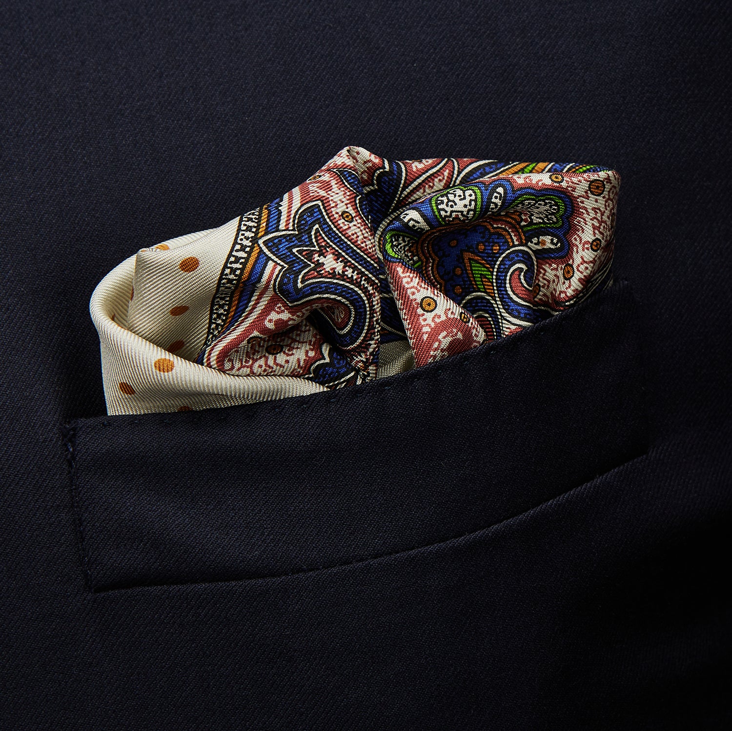View 3 of product The Cream Paisley Polka Dot Silk Pocket Square by Turnbull & Asser features hand-rolled edges, a detailed blue, red, and yellow paisley design with central orange dots, and the brand's name printed in one corner.