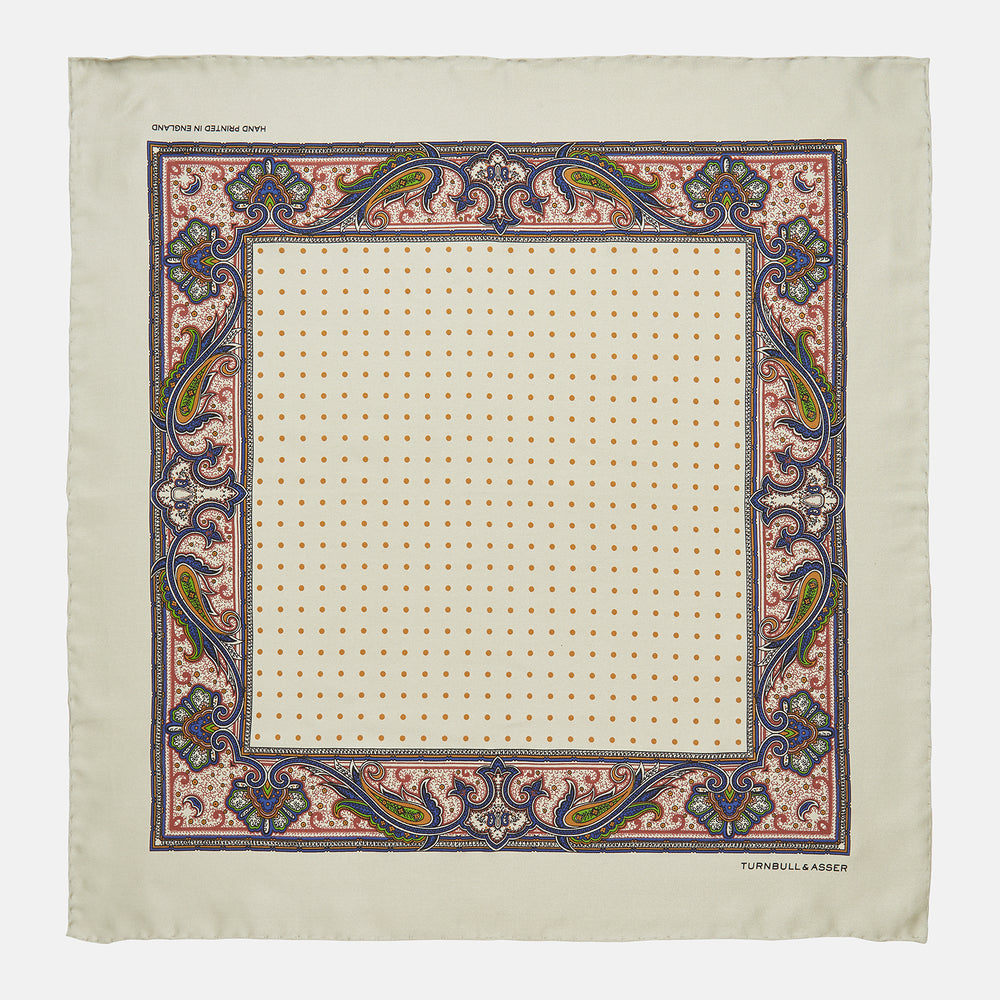 The Cream Paisley Polka Dot Silk Pocket Square by Turnbull & Asser features hand-rolled edges, a detailed blue, red, and yellow paisley design with central orange dots, and the brand's name printed in one corner.