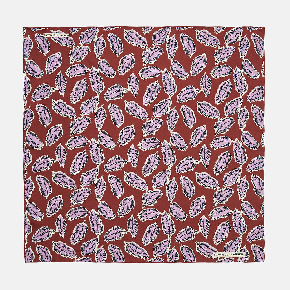 The Red Abstract Feather Print Silk Pocket Square by Turnbull & Asser features a dense purple and black feather pattern on red silk, accented with a discreet brand label—an elegant formal accessory.