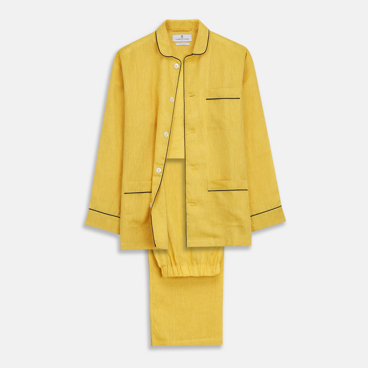 View 2 of product A barefoot person stands against a plain background, wearing the Turnbull & Asser Ochre Linen Modern Pyjama Set with black piping on the collar, cuffs, and pockets. The button-down top completes the look as they gaze to the side.