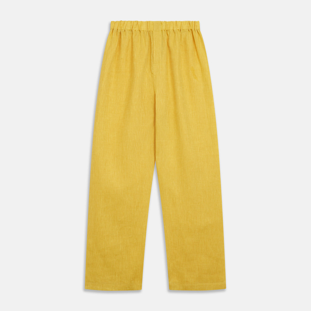 A barefoot person stands against a plain background, wearing the Turnbull & Asser Ochre Linen Modern Pyjama Set with black piping on the collar, cuffs, and pockets. The button-down top completes the look as they gaze to the side.