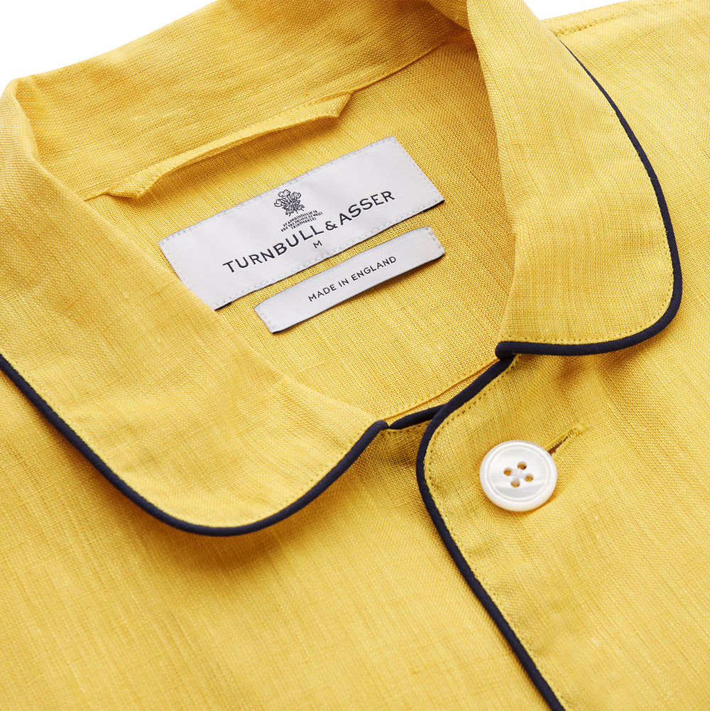 A barefoot person stands against a plain background, wearing the Turnbull & Asser Ochre Linen Modern Pyjama Set with black piping on the collar, cuffs, and pockets. The button-down top completes the look as they gaze to the side.