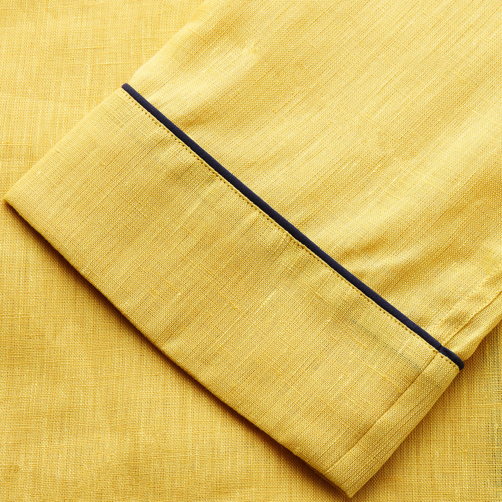 A barefoot person stands against a plain background, wearing the Turnbull & Asser Ochre Linen Modern Pyjama Set with black piping on the collar, cuffs, and pockets. The button-down top completes the look as they gaze to the side.
