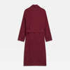 View 6 of product A person stands barefoot against a plain background, wearing the Turnbull & Asser Burgundy Linen Piped Gown belted at the waist over blue pajamas, looking to the side with one hand in the gown’s pocket.