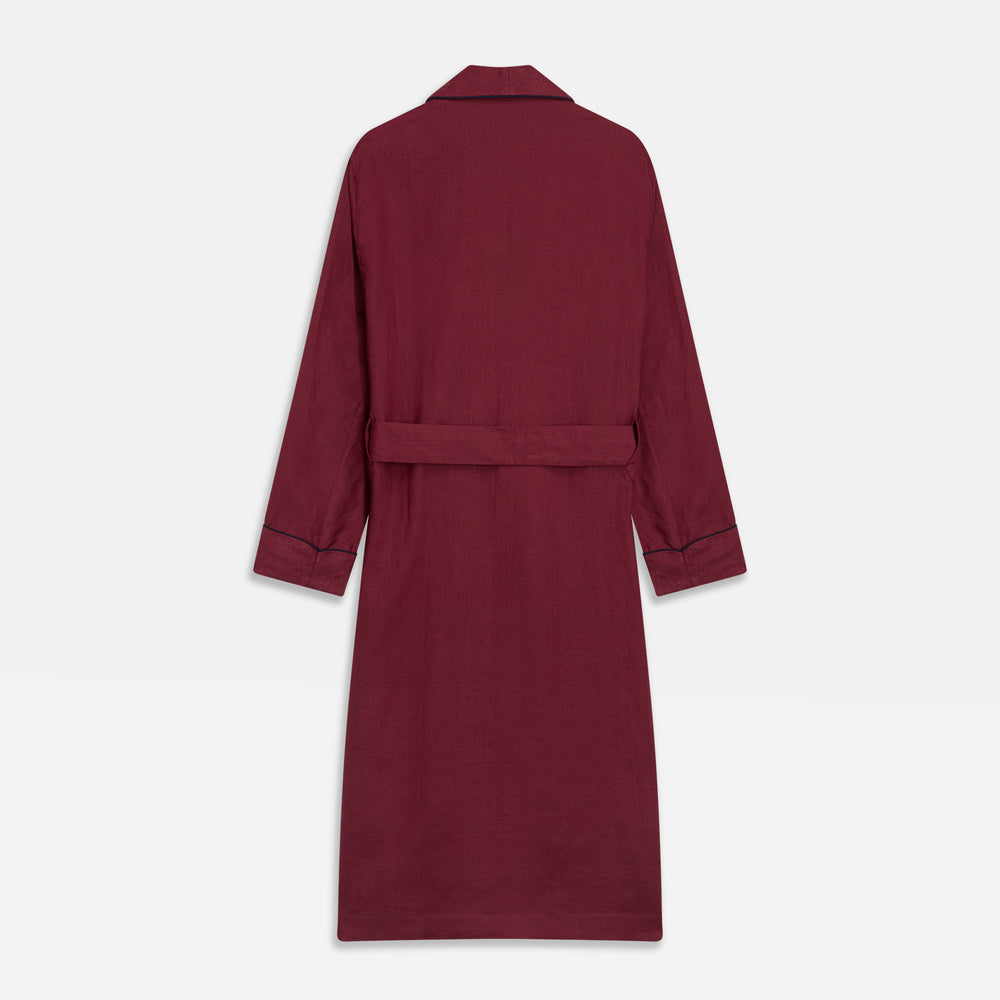 A person stands barefoot against a plain background, wearing the Turnbull & Asser Burgundy Linen Piped Gown belted at the waist over blue pajamas, looking to the side with one hand in the gown’s pocket.