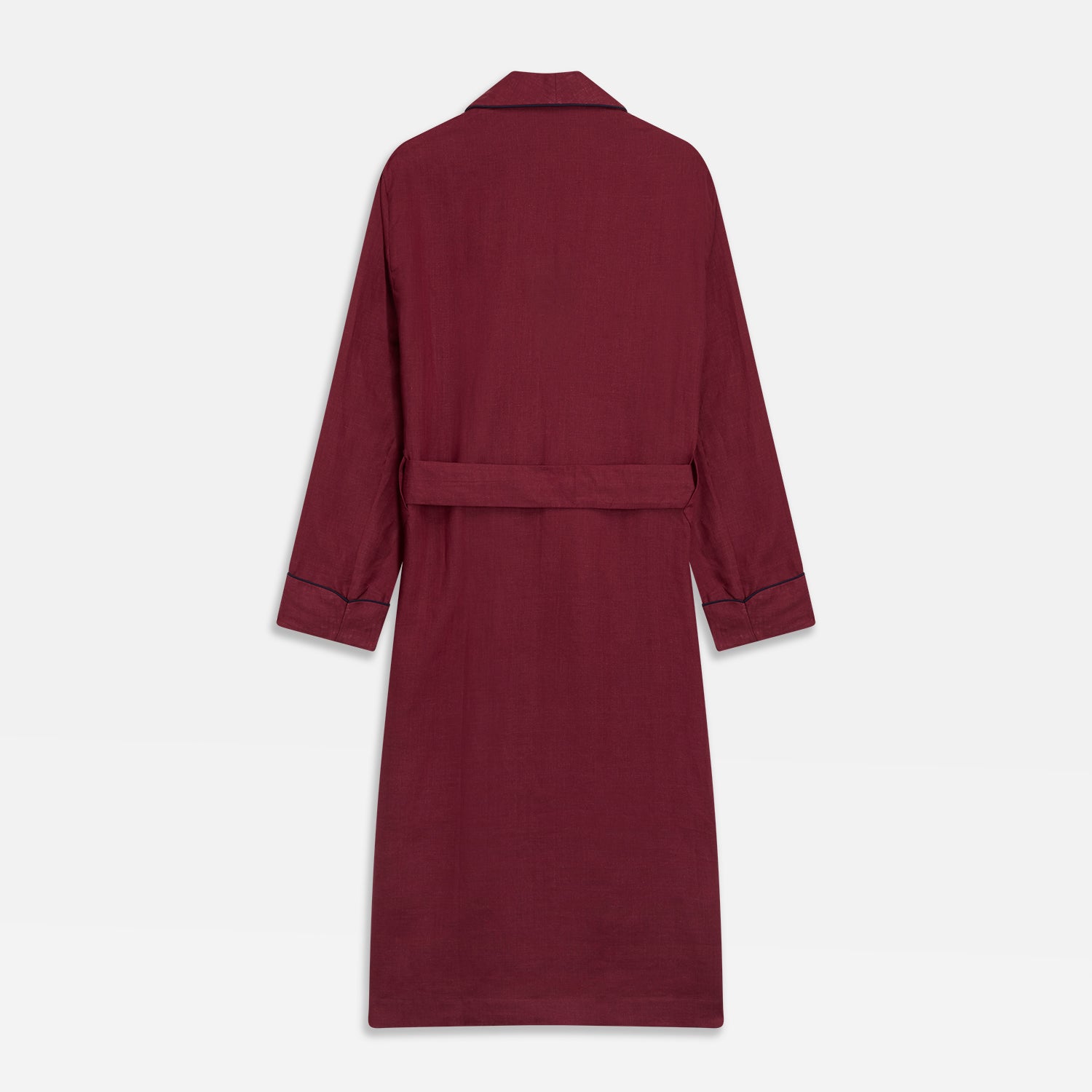 View 6 of product A person stands barefoot against a plain background, wearing the Turnbull & Asser Burgundy Linen Piped Gown belted at the waist over blue pajamas, looking to the side with one hand in the gown’s pocket.
