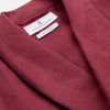 View 4 of product A person stands barefoot against a plain background, wearing the Turnbull & Asser Burgundy Linen Piped Gown belted at the waist over blue pajamas, looking to the side with one hand in the gown’s pocket.