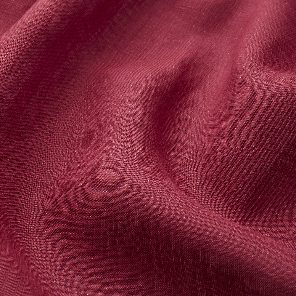 A person stands barefoot against a plain background, wearing the Turnbull & Asser Burgundy Linen Piped Gown belted at the waist over blue pajamas, looking to the side with one hand in the gown’s pocket.