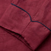View 5 of product A person stands barefoot against a plain background, wearing the Turnbull & Asser Burgundy Linen Piped Gown belted at the waist over blue pajamas, looking to the side with one hand in the gown’s pocket.