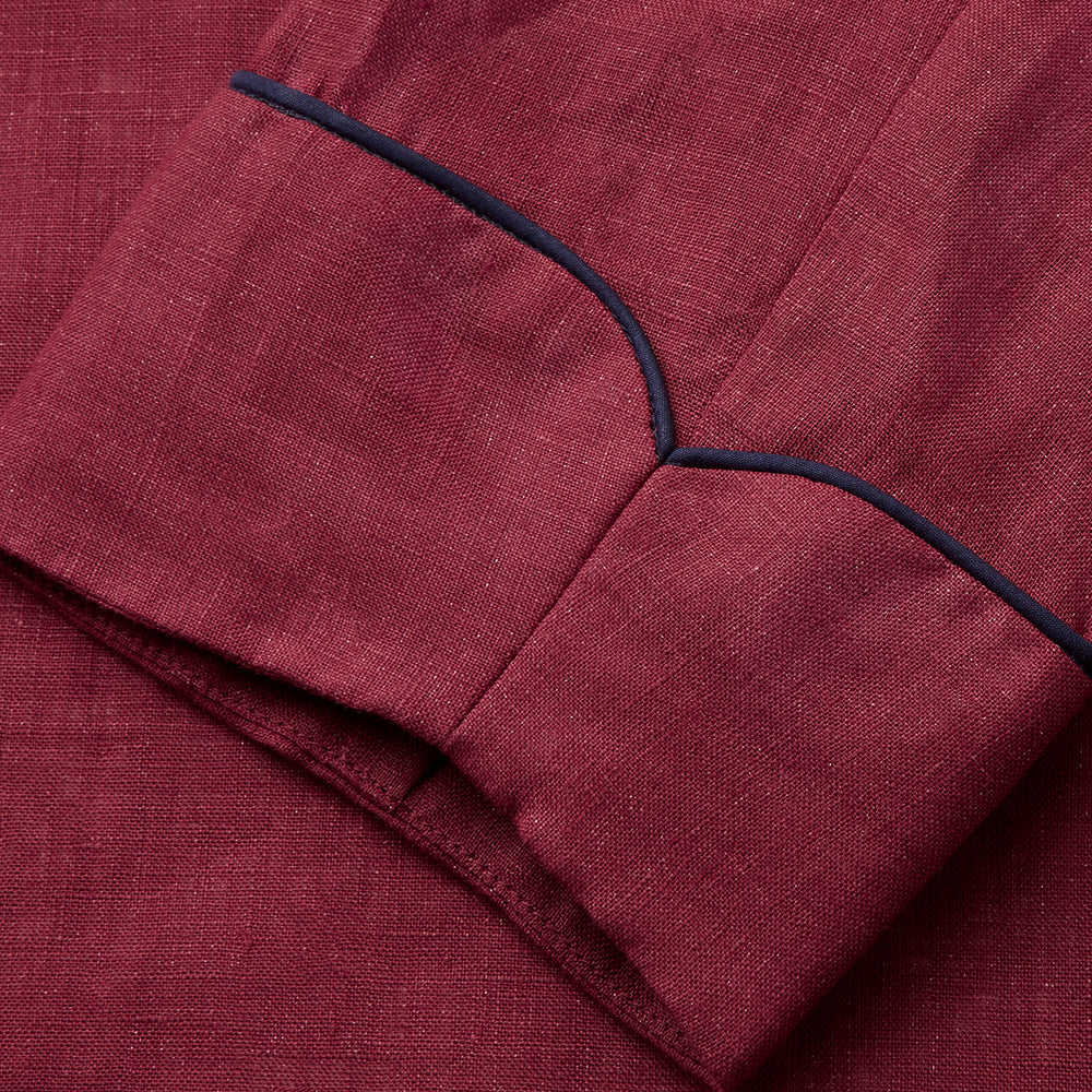 A person stands barefoot against a plain background, wearing the Turnbull & Asser Burgundy Linen Piped Gown belted at the waist over blue pajamas, looking to the side with one hand in the gown’s pocket.