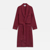 View 2 of product A person stands barefoot against a plain background, wearing the Turnbull & Asser Burgundy Linen Piped Gown belted at the waist over blue pajamas, looking to the side with one hand in the gown’s pocket.