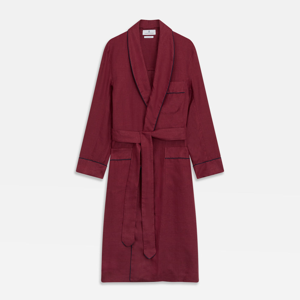 A person stands barefoot against a plain background, wearing the Turnbull & Asser Burgundy Linen Piped Gown belted at the waist over blue pajamas, looking to the side with one hand in the gown’s pocket.