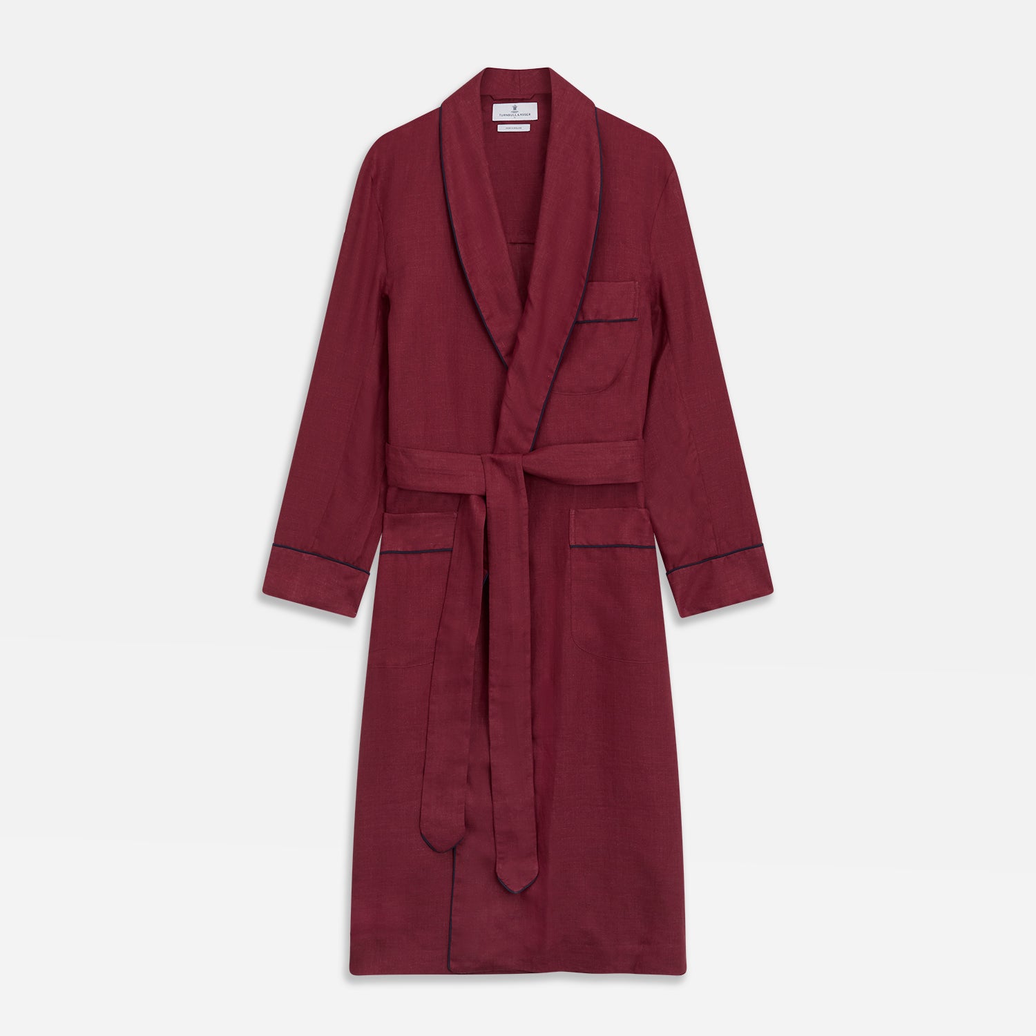 View 2 of product A person stands barefoot against a plain background, wearing the Turnbull & Asser Burgundy Linen Piped Gown belted at the waist over blue pajamas, looking to the side with one hand in the gown’s pocket.