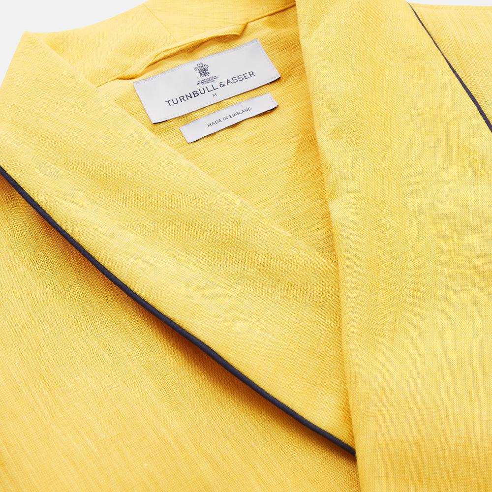 The Turnbull & Asser Ochre Linen Piped Gown features black piping on the collar, cuffs, and edges, a chest pocket, two front pockets, and a matching waist belt. Made with breathable linen for comfort, shown against a plain white background.