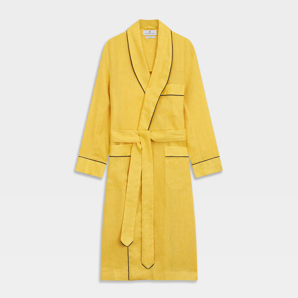 The Turnbull & Asser Ochre Linen Piped Gown features black piping on the collar, cuffs, and edges, a chest pocket, two front pockets, and a matching waist belt. Made with breathable linen for comfort, shown against a plain white background.