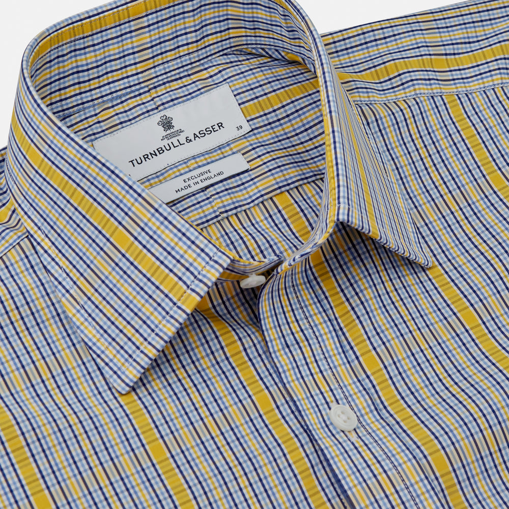 A man stands against a light background, wearing a Turnbull & Asser Yellow and Blue Fancy Check Regular Fit Shirt with T&A Collar, a patterned tie, dark dress pants, and black shoes. He faces forward with hands in pockets and a neutral expression.