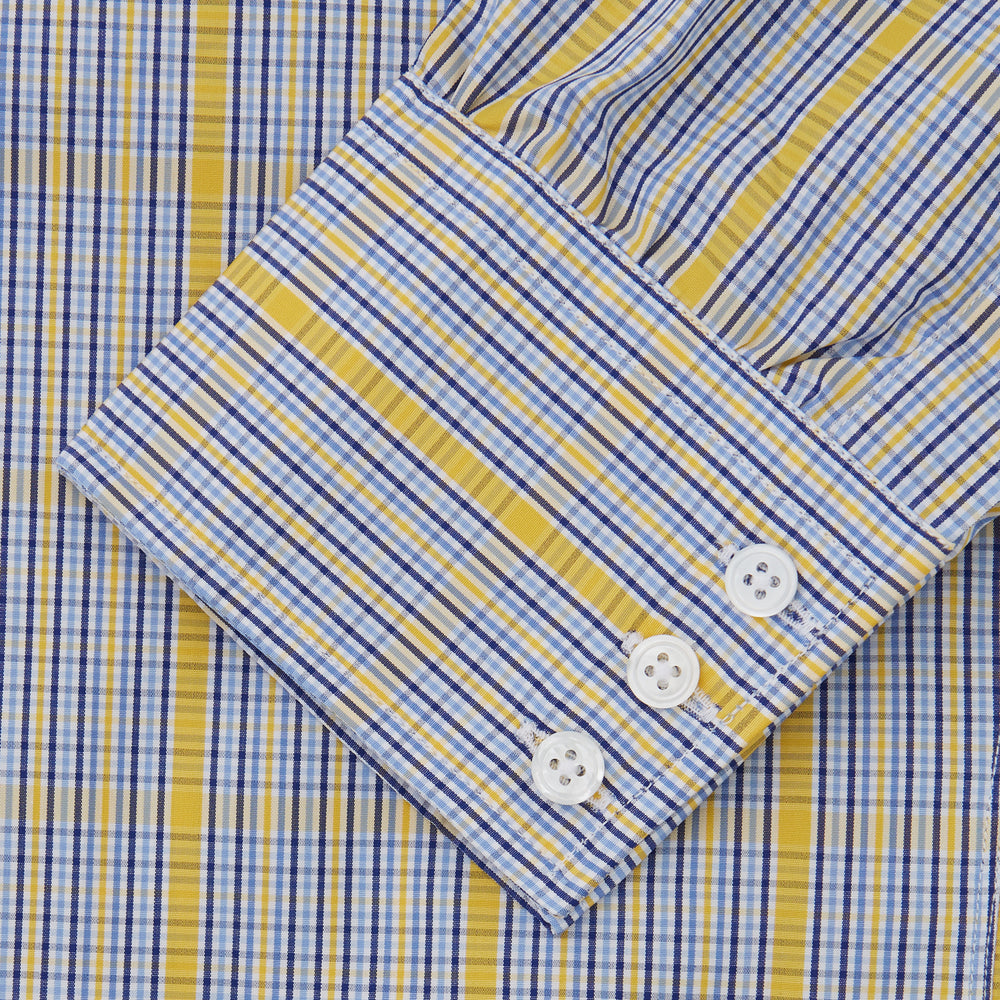 A man stands against a light background, wearing a Turnbull & Asser Yellow and Blue Fancy Check Regular Fit Shirt with T&A Collar, a patterned tie, dark dress pants, and black shoes. He faces forward with hands in pockets and a neutral expression.