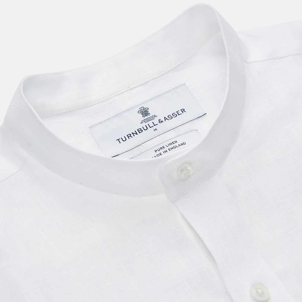 A man stands against a beige background, wearing the Turnbull & Asser White Linen Weekend Fit Blake Shirt, dark trousers, and beige sneakers. With one hand in his pocket and a neutral expression, he captures effortless weekend style.