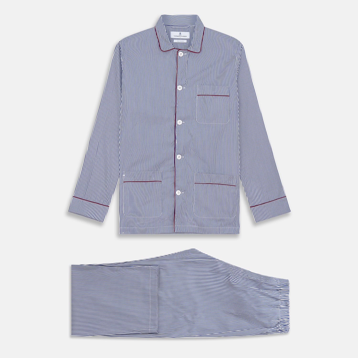 View 1 of product The Turnbull & Asser Navy & White Stripe Cotton Modern Pyjama Set features blue and white Bengal stripes with red piping, a long-sleeve button-up top, chest pocket, and matching pants. Shown laid flat on a white background.