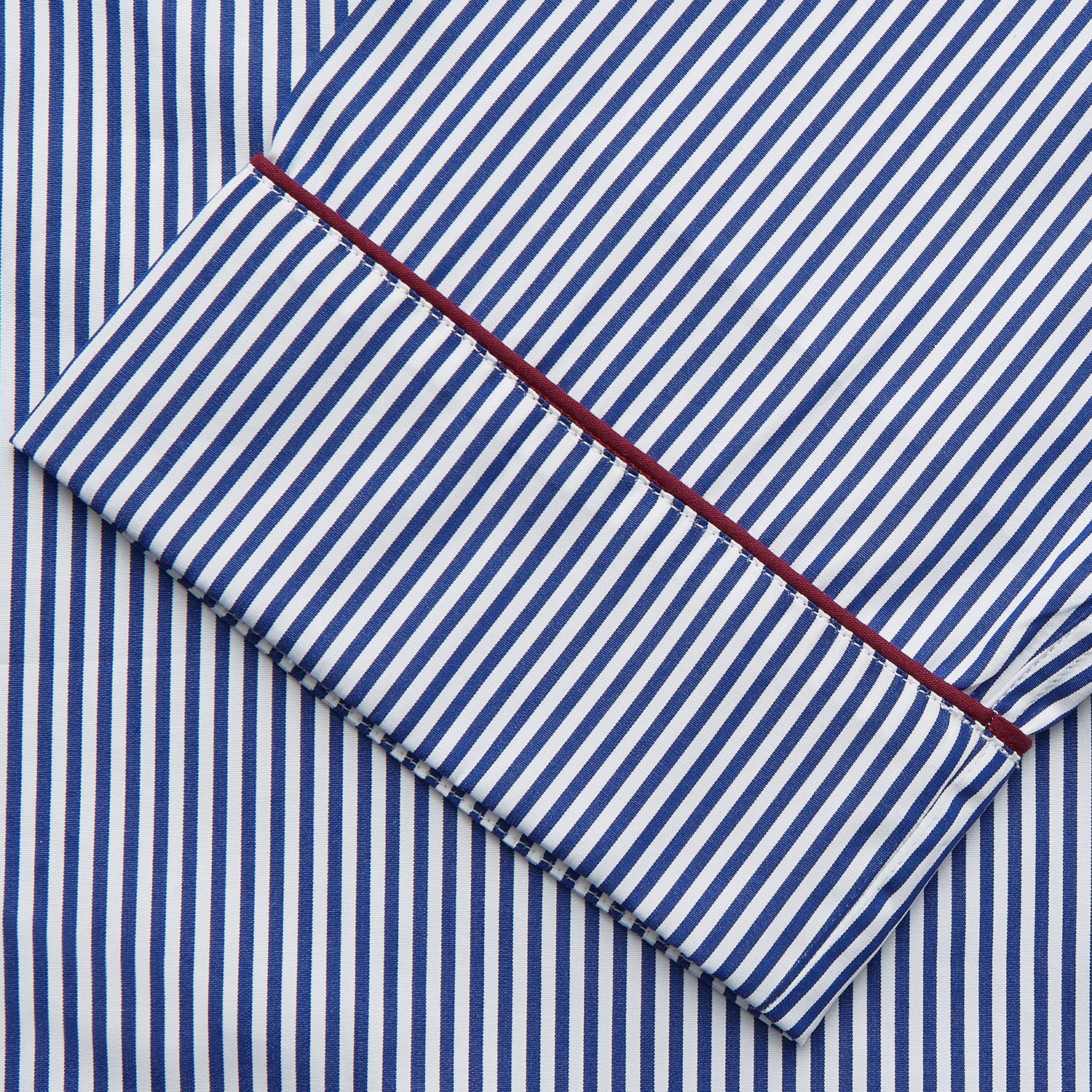 View 2 of product The Turnbull & Asser Navy & White Stripe Cotton Modern Pyjama Set features blue and white Bengal stripes with red piping, a long-sleeve button-up top, chest pocket, and matching pants. Shown laid flat on a white background.