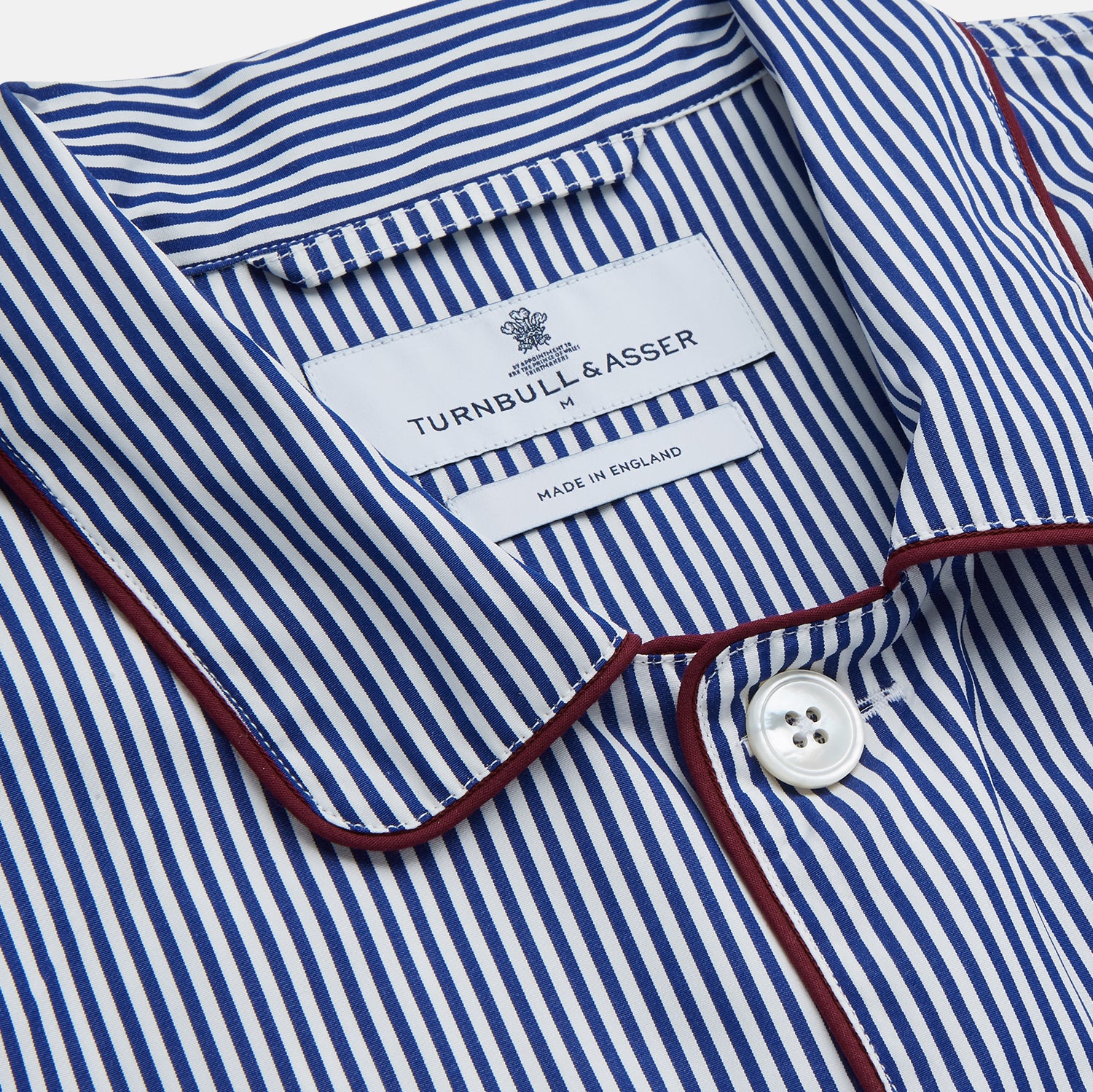 View 3 of product The Turnbull & Asser Navy & White Stripe Cotton Modern Pyjama Set features blue and white Bengal stripes with red piping, a long-sleeve button-up top, chest pocket, and matching pants. Shown laid flat on a white background.