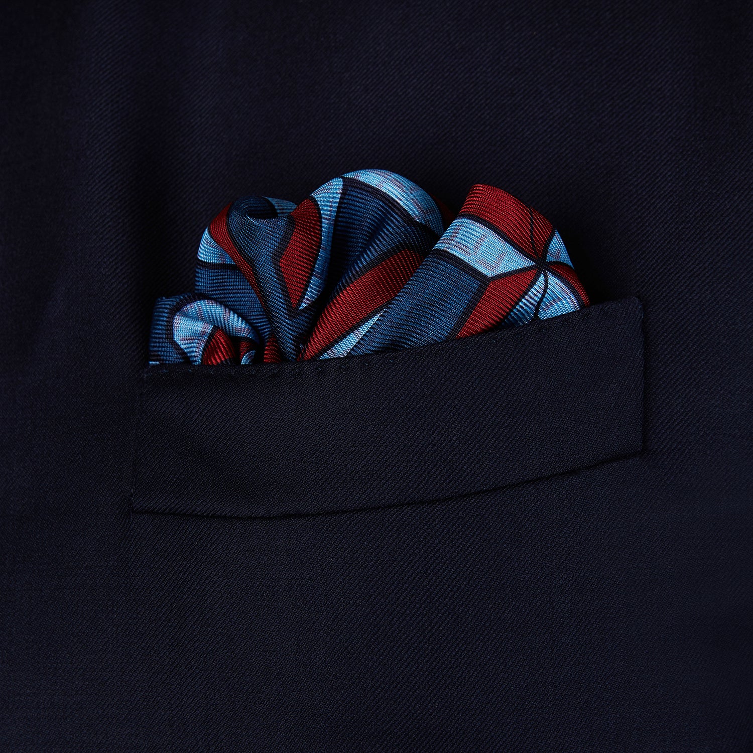 View 3 of product The Turnbull & Asser Red Multi Crosses Double-Faced Silk Pocket Square features red, white, and light blue Xs on dark blue silk with an orange border and the brand's name printed in the bottom right corner.