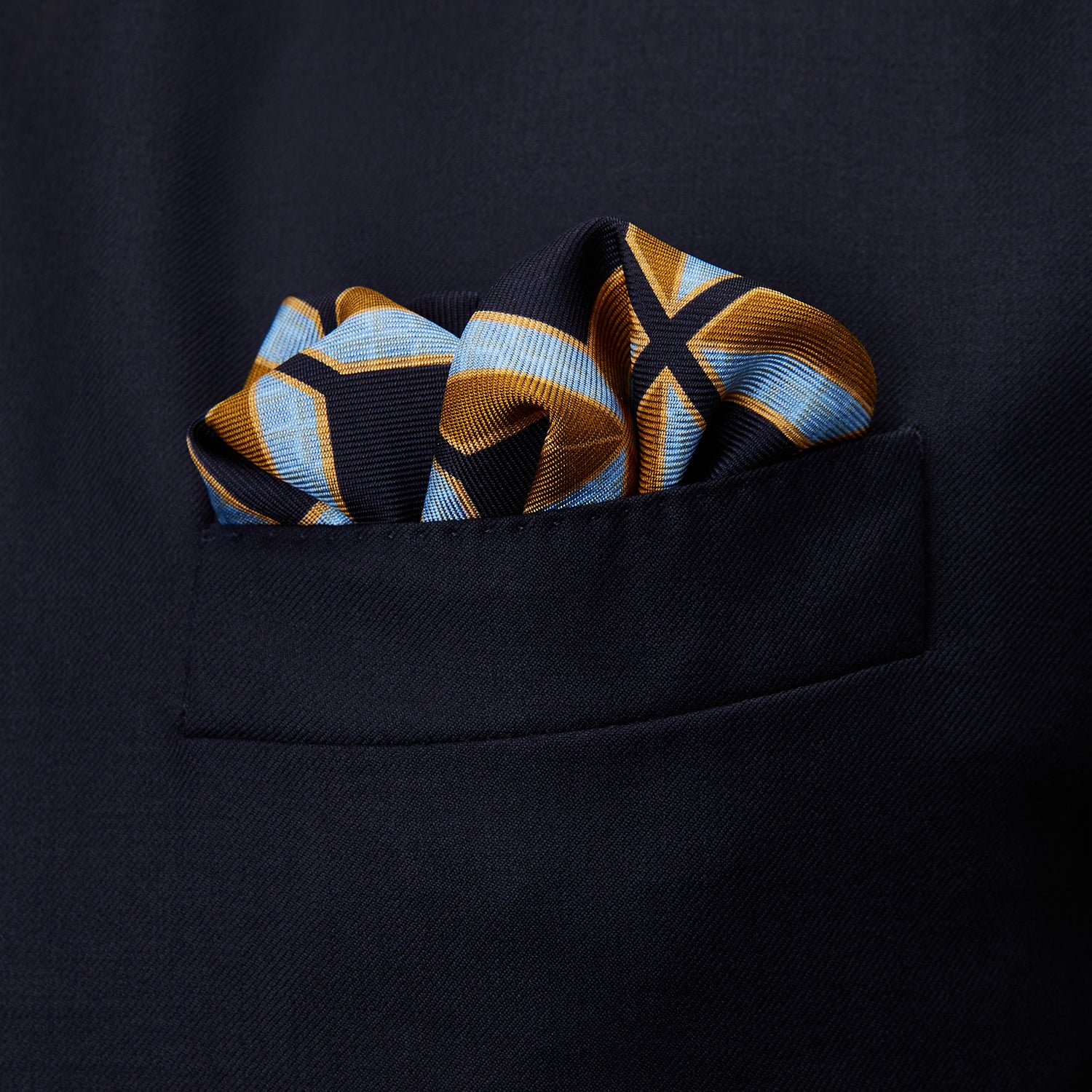 View 3 of product The Turnbull & Asser Navy Multi Crosses Double-Faced Silk Pocket Square features intersecting yellow and white X-shapes on a dark background with a red border, and “TURNBULL & ASSER” plus “PRINTED IN ENGLAND” in two corners.