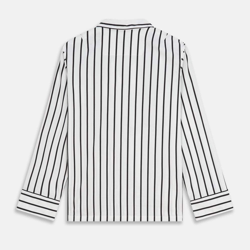 A person stands barefoot on a concrete floor, wearing the Turnbull & Asser Black & White Stripe Harriet Women’s Pyjama Set, featuring loose-fitting white pajamas with black vertical stripes, against a plain beige wall in soft side lighting.