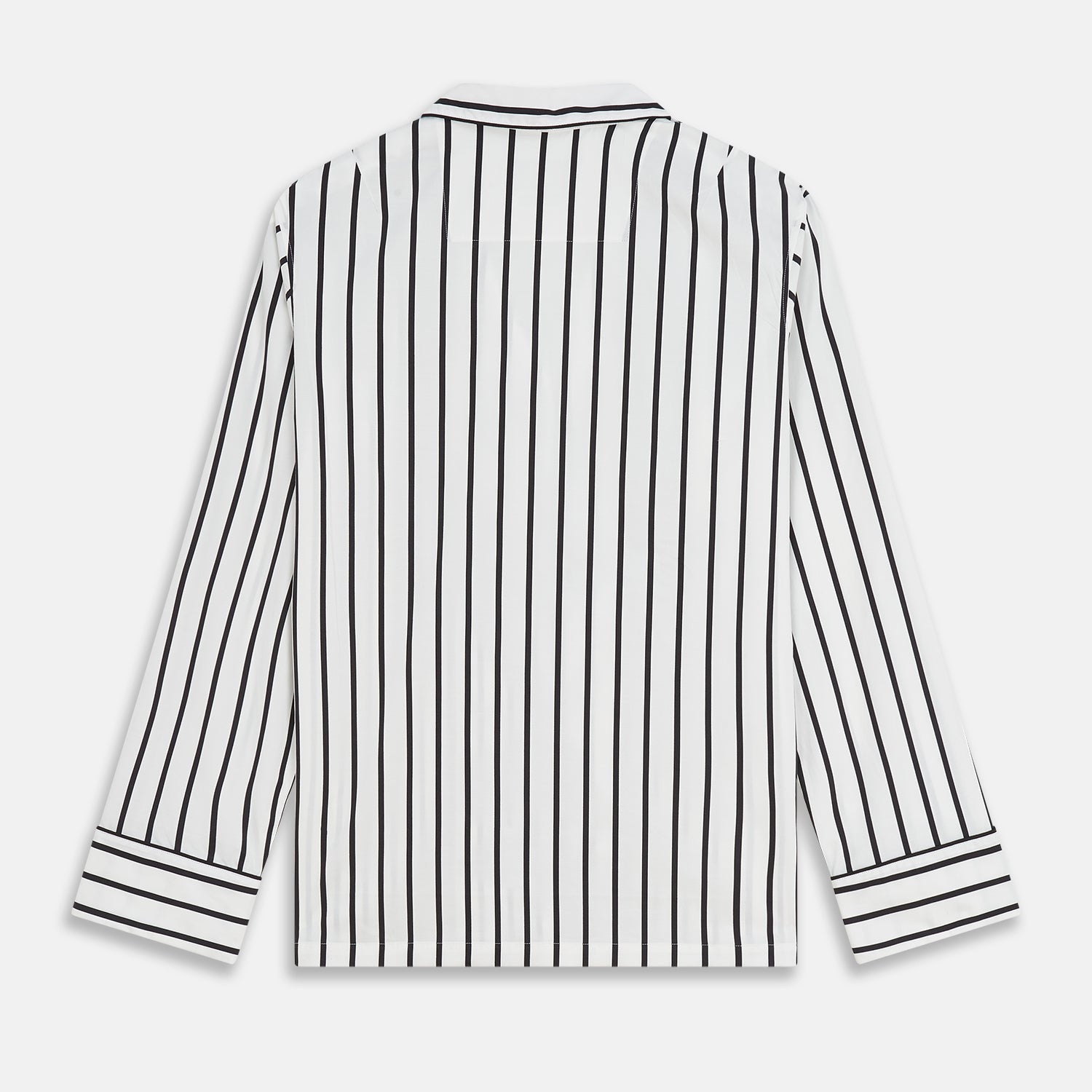 View 6 of product A person stands barefoot on a concrete floor, wearing the Turnbull & Asser Black & White Stripe Harriet Women’s Pyjama Set, featuring loose-fitting white pajamas with black vertical stripes, against a plain beige wall in soft side lighting.
