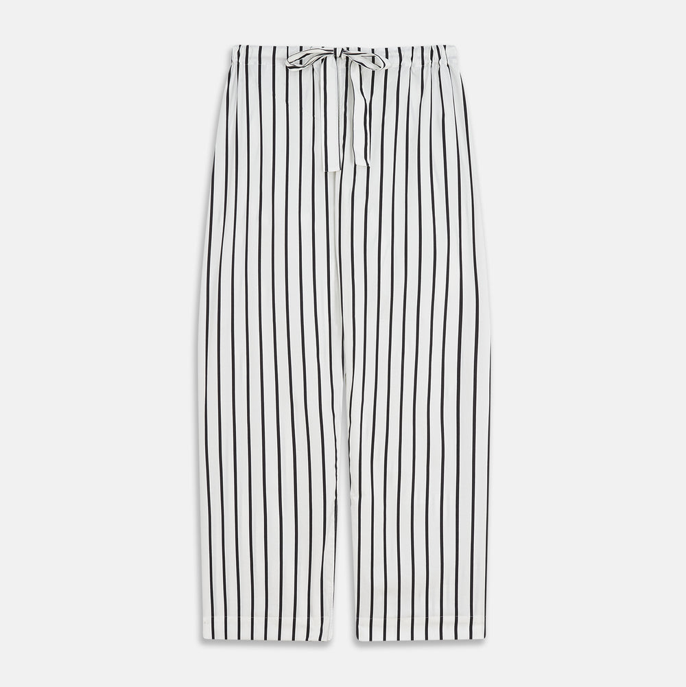 A person stands barefoot on a concrete floor, wearing the Turnbull & Asser Black & White Stripe Harriet Women’s Pyjama Set, featuring loose-fitting white pajamas with black vertical stripes, against a plain beige wall in soft side lighting.