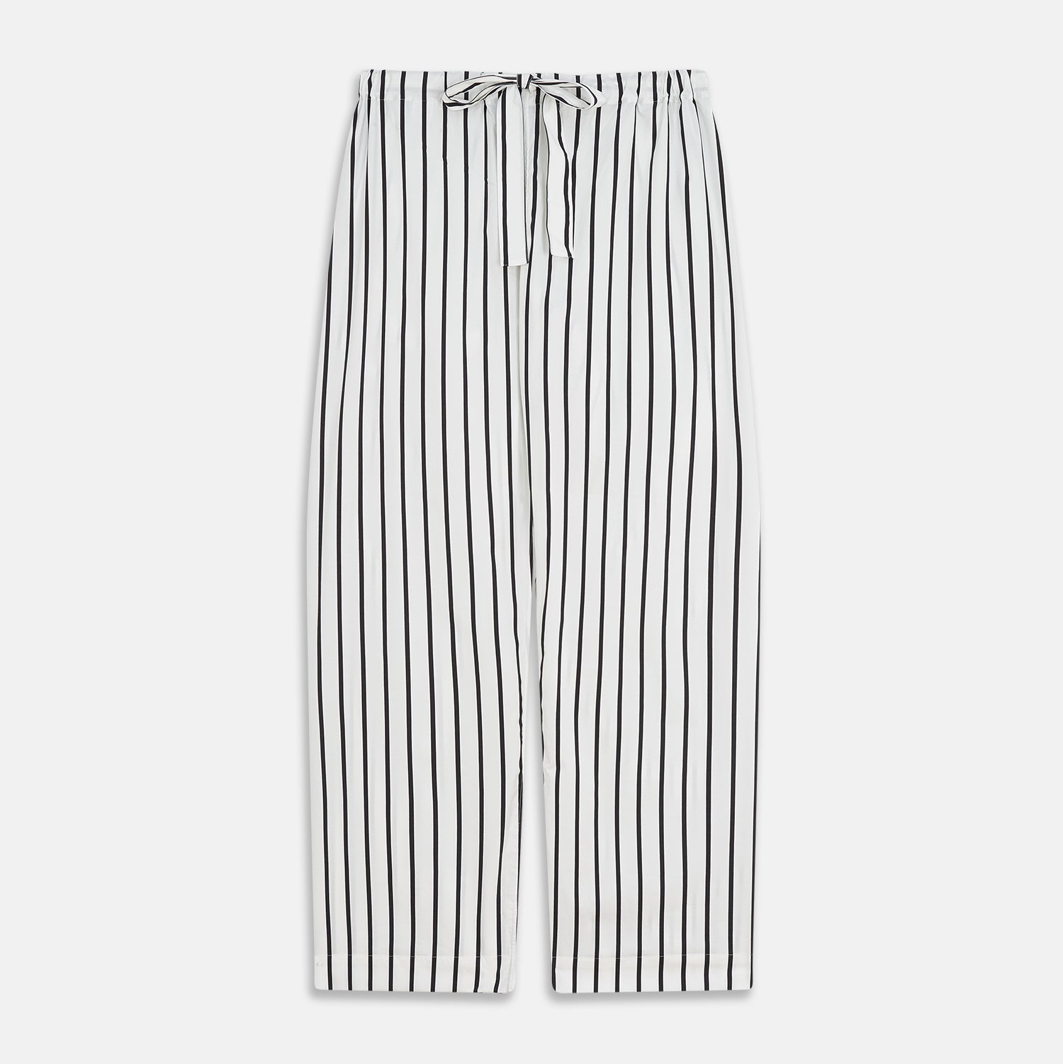 View 7 of product A person stands barefoot on a concrete floor, wearing the Turnbull & Asser Black & White Stripe Harriet Women’s Pyjama Set, featuring loose-fitting white pajamas with black vertical stripes, against a plain beige wall in soft side lighting.