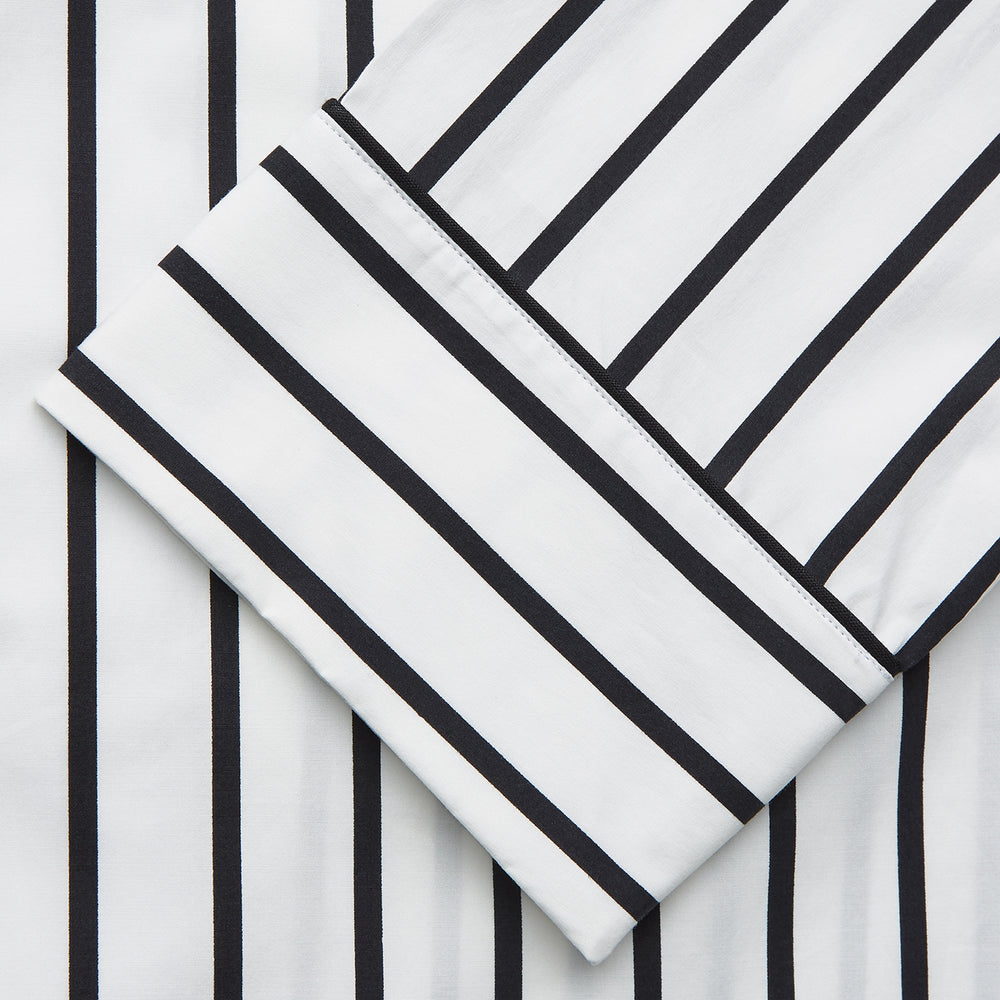 A person stands barefoot on a concrete floor, wearing the Turnbull & Asser Black & White Stripe Harriet Women’s Pyjama Set, featuring loose-fitting white pajamas with black vertical stripes, against a plain beige wall in soft side lighting.