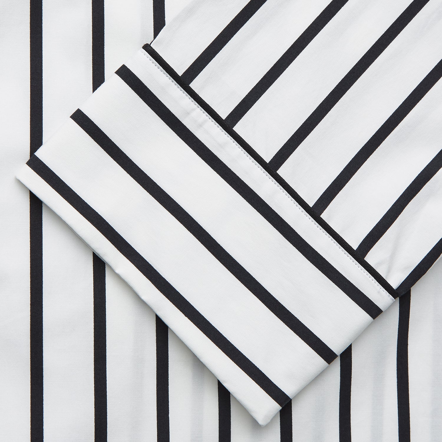 View 3 of product A person stands barefoot on a concrete floor, wearing the Turnbull & Asser Black & White Stripe Harriet Women’s Pyjama Set, featuring loose-fitting white pajamas with black vertical stripes, against a plain beige wall in soft side lighting.