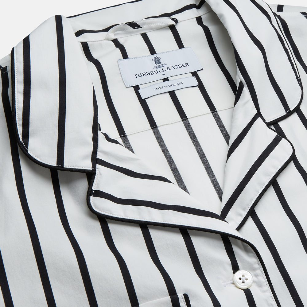 A person stands barefoot on a concrete floor, wearing the Turnbull & Asser Black & White Stripe Harriet Women’s Pyjama Set, featuring loose-fitting white pajamas with black vertical stripes, against a plain beige wall in soft side lighting.