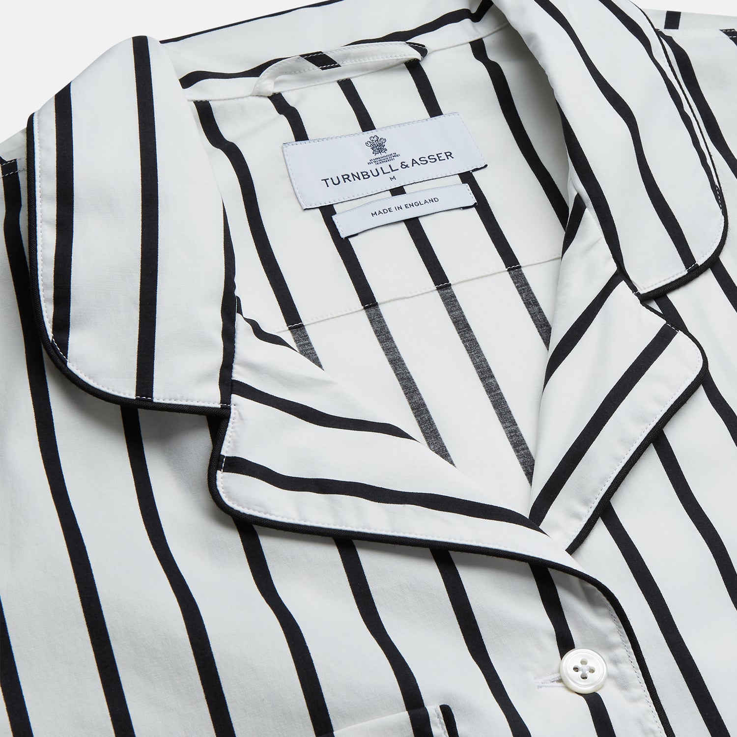 View 4 of product A person stands barefoot on a concrete floor, wearing the Turnbull & Asser Black & White Stripe Harriet Women’s Pyjama Set, featuring loose-fitting white pajamas with black vertical stripes, against a plain beige wall in soft side lighting.