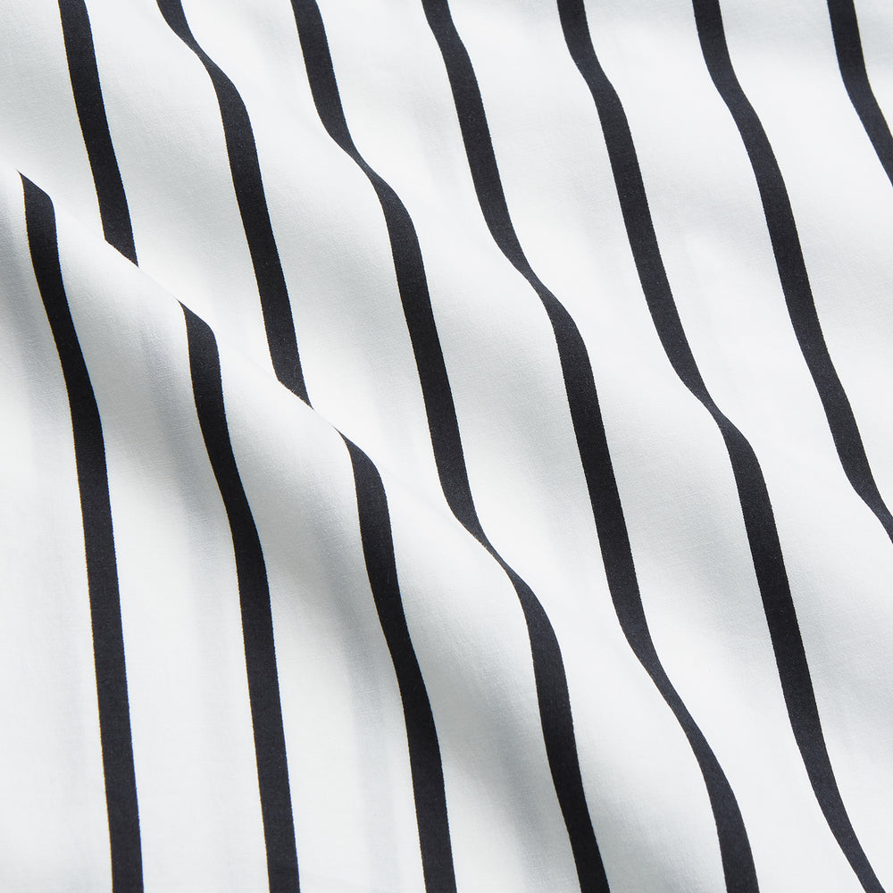 A person stands barefoot on a concrete floor, wearing the Turnbull & Asser Black & White Stripe Harriet Women’s Pyjama Set, featuring loose-fitting white pajamas with black vertical stripes, against a plain beige wall in soft side lighting.