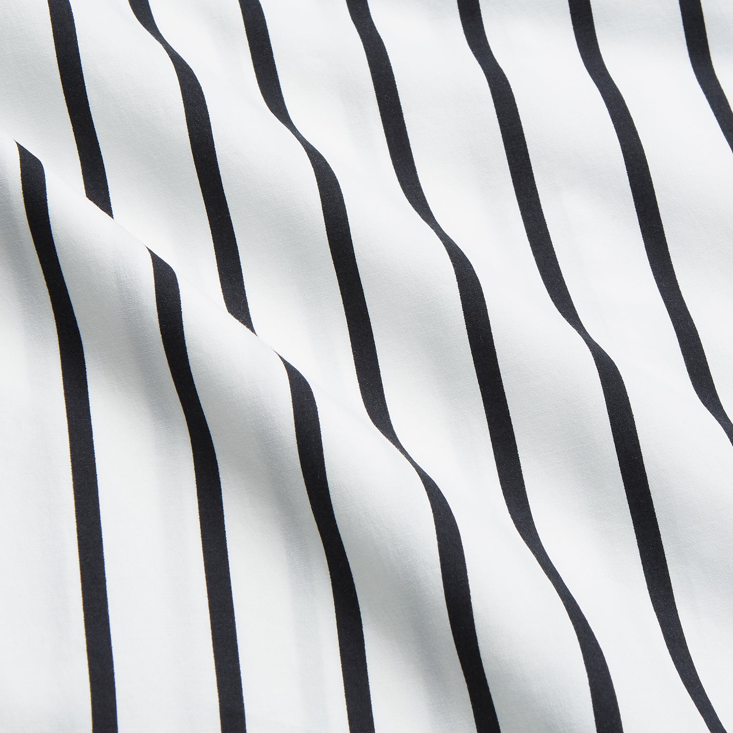 View 5 of product A person stands barefoot on a concrete floor, wearing the Turnbull & Asser Black & White Stripe Harriet Women’s Pyjama Set, featuring loose-fitting white pajamas with black vertical stripes, against a plain beige wall in soft side lighting.