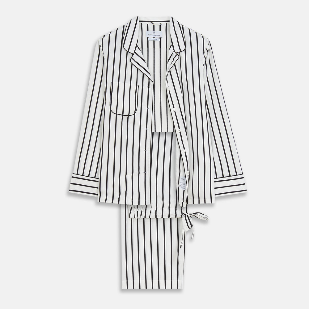 A person stands barefoot on a concrete floor, wearing the Turnbull & Asser Black & White Stripe Harriet Women’s Pyjama Set, featuring loose-fitting white pajamas with black vertical stripes, against a plain beige wall in soft side lighting.