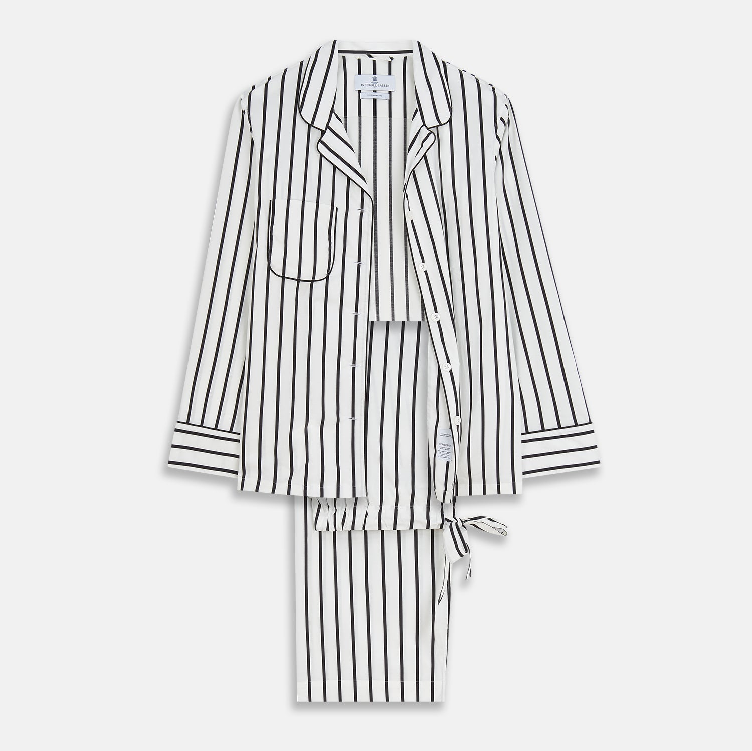 View 2 of product A person stands barefoot on a concrete floor, wearing the Turnbull & Asser Black & White Stripe Harriet Women’s Pyjama Set, featuring loose-fitting white pajamas with black vertical stripes, against a plain beige wall in soft side lighting.