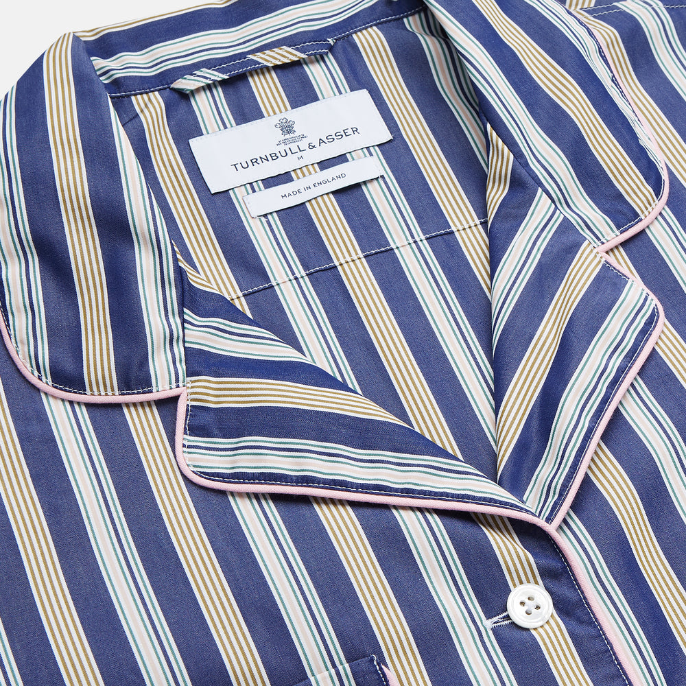 A person stands barefoot on a gray floor in the Turnbull & Asser Blue Multi Stripe Harriet Women’s Pyjama Set with a dark sweater over their shoulders—premium cotton pyjamas that make a perfect gift for her—against a plain beige background.