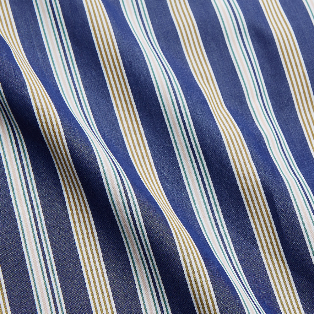 A person stands barefoot on a gray floor in the Turnbull & Asser Blue Multi Stripe Harriet Women’s Pyjama Set with a dark sweater over their shoulders—premium cotton pyjamas that make a perfect gift for her—against a plain beige background.