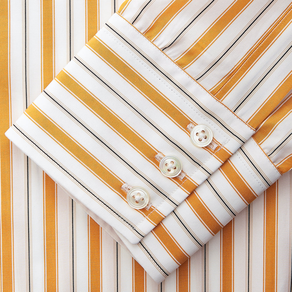 A man stands against a tan background wearing a Turnbull & Asser Orange Multi Stripe Regular Fit Shirt with T&A Collar and 3 Button Cuffs, paired with tan dress pants, brown belt, and brown shoes. He faces forward with a neutral expression.