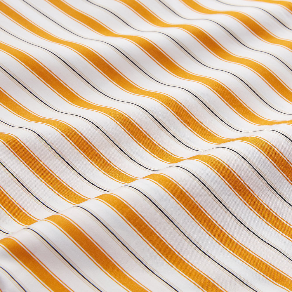 A man stands against a tan background wearing a Turnbull & Asser Orange Multi Stripe Regular Fit Shirt with T&A Collar and 3 Button Cuffs, paired with tan dress pants, brown belt, and brown shoes. He faces forward with a neutral expression.
