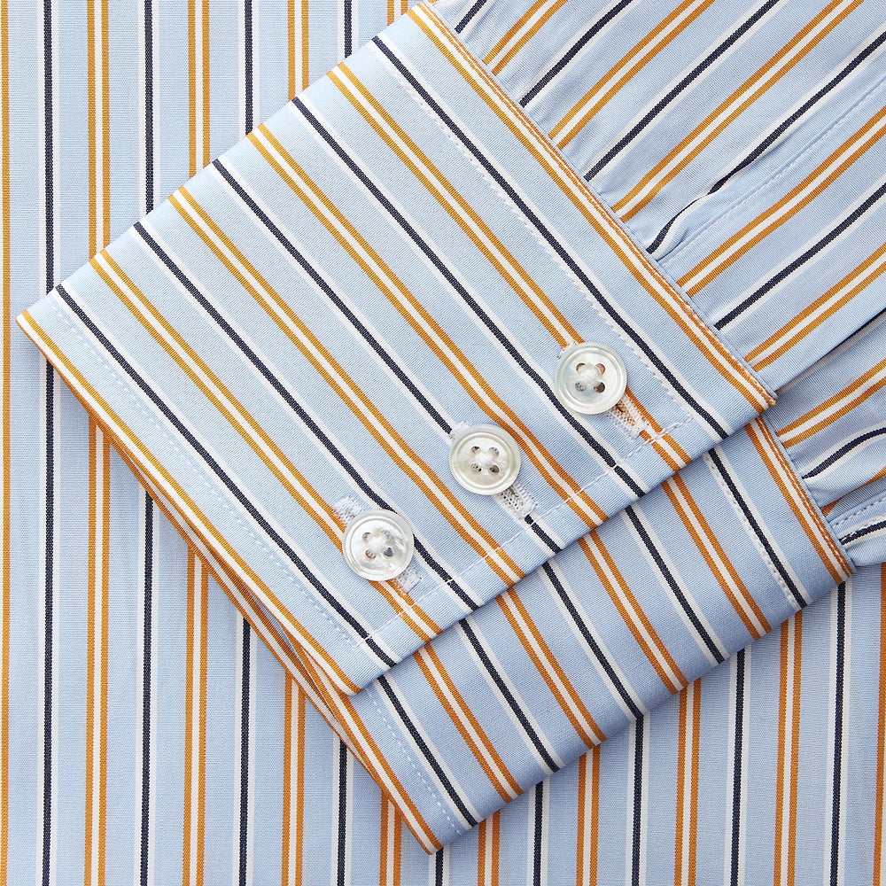 A man stands against a beige background, wearing a Turnbull & Asser Blue, Orange & Navy Multi Stripe Regular Fit Shirt with T&A collar and 3-button cuffs, tucked into tan dress pants with a black belt. He faces forward with a neutral expression.